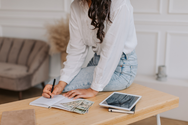 how to be financially free: a woman in a shirt and jeans writing in a notebook creating a budget