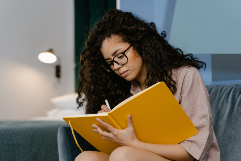 woman with curly hair and glasses writing in a yellow journal
